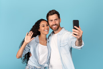Cheerful young caucasian man with beard hugs arabic lady, waving hand, have video call on smartphone