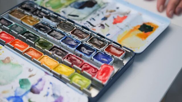 Closeup view of colorful watercolor palette on table with paintbrushes and painter tools. Woman hands holding paintbrush and drawing on white paper canvas with aquarelle