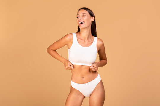 Portrait of slim smiling model woman in underwear top bra and panties standing isolated over beige studio background