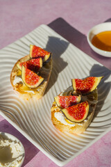 Open sandwiches or bruschetta with cream cheese, fresh figs, honey and walnuts on a white rectangular plate on a pink concrete background.