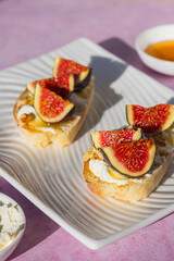 Open sandwiches or bruschetta with cream cheese, fresh figs, honey and walnuts on a white rectangular plate on a pink concrete background.