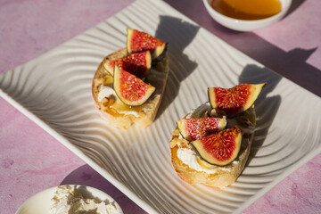 Open sandwiches or bruschetta with cream cheese, fresh figs, honey and walnuts on a white rectangular plate on a pink concrete background.
