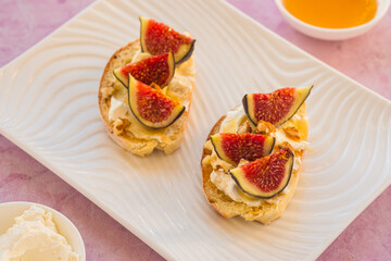 Open sandwiches or bruschetta with cream cheese, fresh figs, honey and walnuts on a white rectangular plate on a pink concrete background.