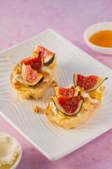 Open sandwiches or bruschetta with cream cheese, fresh figs, honey and walnuts on a white rectangular plate on a pink concrete background.