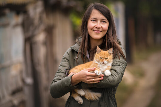 Portrait of young woman glasses holding cute red cat with green eyes. Female hugging her cute long hair kitty. Background, copy space, outdoors beautiful playing brunette.