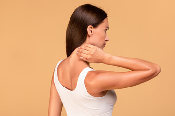 Young woman touching and self massaging her neck, rubbing skin, standing over beige studio background, free space