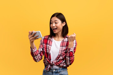 Overjoyed asian lady holding modern smartphone and clenching fist, gambling or trading online over yellow background