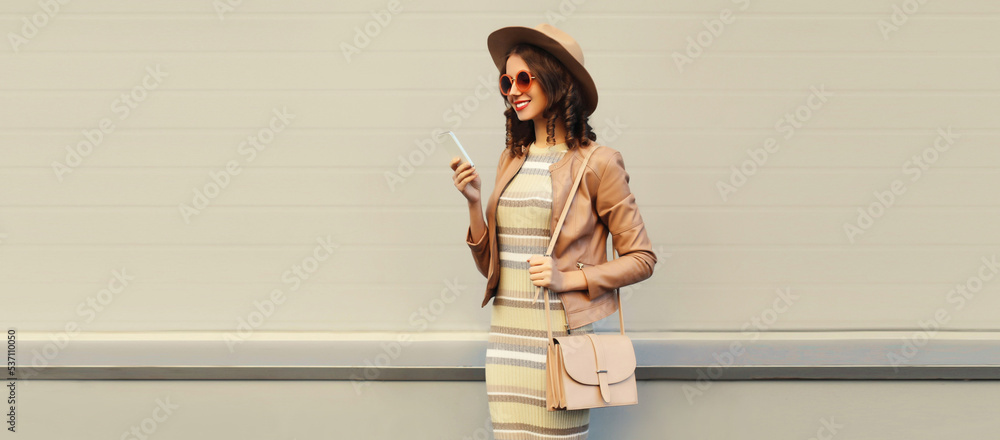 Wall mural portrait of stylish smiling woman with smartphone wearing round hat, leather jacket on gray backgrou