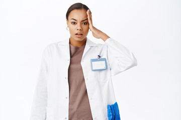 Troubled female doctor, holds hand on head, look concerned and complicated, stands against white background