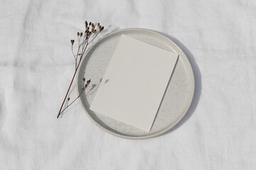 Blank greeting card card mockup on ceramic plate, tray. Dry grass, plant in sunlight. White linen...