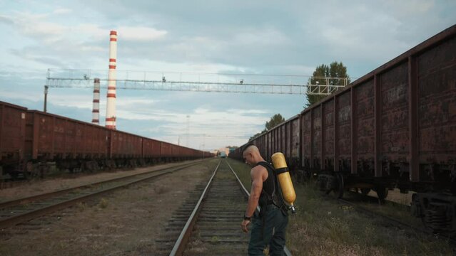 Young muscular man with a balloon drager on his back is training on the railway sidings next to a factory with chimneys