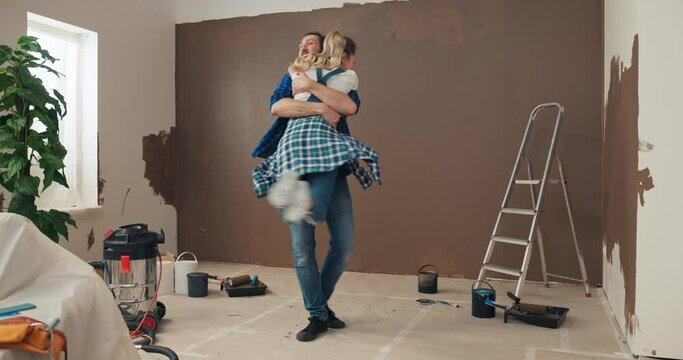 Young man and woman are renovating new apartment. They are full of emotions, happy, hugging and laughing. The husband raised wife in arms and spins. They admire new home. There is ladder behind.