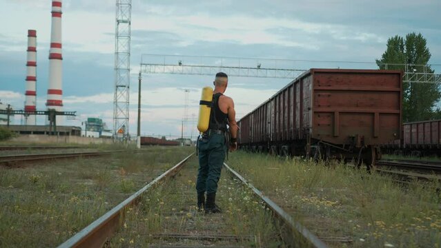 Bodybuilder with balloon drager on his back stands on the rails near the factory and rests between exercises. Back view