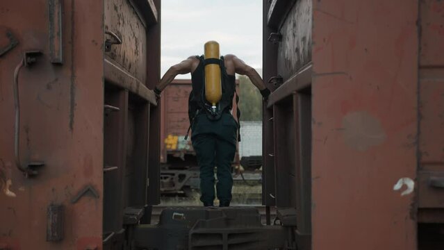 Bodybuilder with balloon drager on his back pulls himself up on his hands between freight wagons. Street workout. Back view