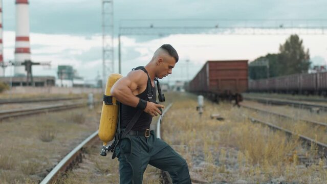 Young muscular man stands on the rails of a marshalling yard and puts on balloon dragger on his back. Street workout