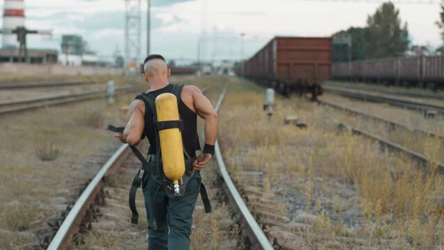 Young muscular man goes along the rails of marshalling yard and removes a drager compressed air tank from his back
