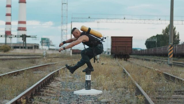 Bodybuilder with balloon drager on his back training at railway yard near factory and squats on one leg standing on column