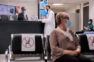 Male health care professional preparing patient records for care at hospital clinic. Medical staff, several cultures wearing masks patients protecting themselves from covid 19 at sanatorium lobby. - obrazy, fototapety, plakaty
