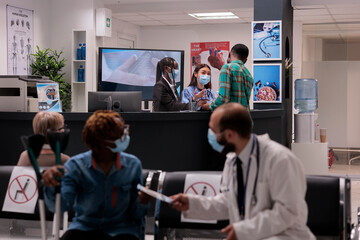 Obraz premium Female hospital staff members attending a sick young african american man at reception area. Doctor handing prescription to woman with partial body paralysis. Medical clinic lobby during covid