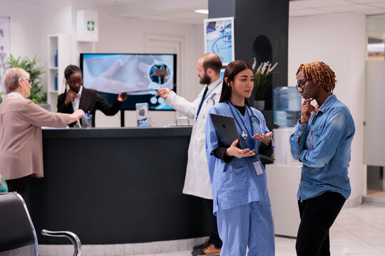 Asian female medical staff sharing on laptop screen clinical images with african american patient at hospital reception. Young receptionist attending doctor, elderly woman at desk.