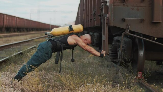 Young muscular man with balloon drager on his back is exercising at railway yard and doing push-ups on the steps of freight wagon