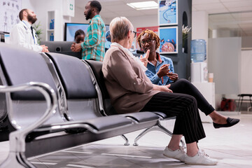 Women talking each other at hospital waiting area. African american man talking to general practitioner. People awaiting in sanatorium admissions section. Elderly lady explaining condition to girl. - obrazy, fototapety, plakaty