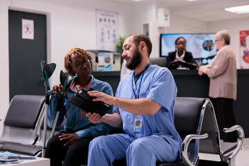 African american woman looking images of injuries on tablet. Physician at rehabilitation clinic explaining therapies to young patient with leg conditions. Sanatorium waiting room with diverse people. - obrazy, fototapety, plakaty