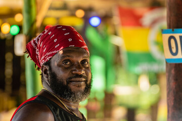 Retrato horizontal de cerca del rostro de un hombre afrocaribeño de barba con un pañuelo rojo en su cabeza al aire libre sonriendo mirando a cámara. 