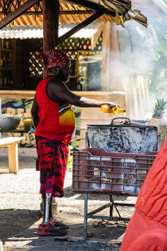 Imagen vertical de un hombre afrocaribeño de barba con una prótesis en su pierna y un pañuelo rojo en su cabeza, de pie al aire libre cocinando en su carne a la parrilla. 