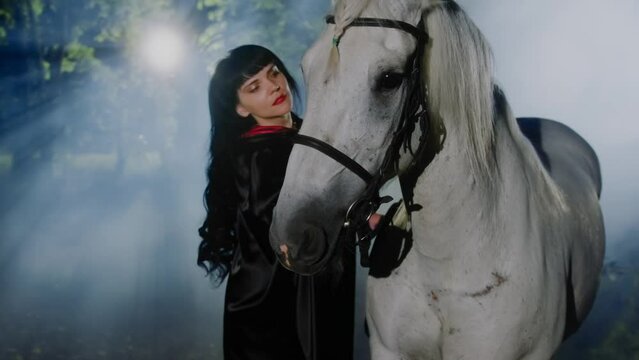 A fantasy girl sorceress soothes a horse by stroking its mane while standing in a foggy fantasy forest at night. The horse is worried. 