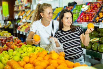 Obraz premium Young woman with a fifteen-year-old girl who came to the supermarket for shopping, choose oranges at the counter, putting ..them in a package