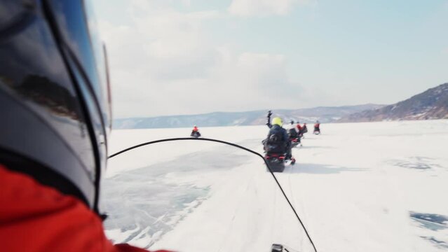 Shooting from the first person. Many snowmobiles ride in a row on snow-covered ice against the backdrop of mountains. Entertainment on Lake Baikal in winter. Tourist leisure. Very cold weather.