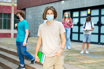 Teenager boy in face mask going home after classes in high school. Other teens walking in background and wearing face masks too.