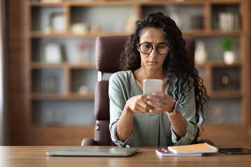Concentrated woman entrepreneur sitting at workplace, using smartphone