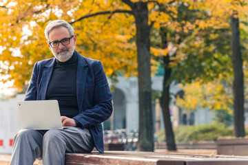Elegant caucasian gray-haired man working remotely on his white laptop from park bench. Office break. Golden autumn. Sunny weather. Yellow and orange leaves. High quality photo