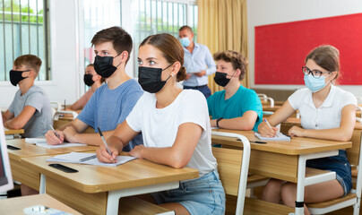 Obraz na płótnie Canvas Teenagers in protective mask listening to lecturer and writing in notebooks in classroom