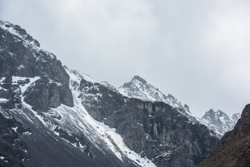 Obraz premium Awesome landscape with high snowy mountain peaked top with sharp rocks in cloudy sky. Dramatic view to snow mountain pointed peak in rainy weather. Atmospheric scenery with white snow on black rocks.
