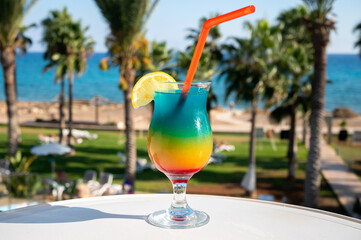 Colourful cold Rainbow Paradise cocktail drink served in glass at pool bar overlooking blue pool, sea and palm trees, relax and holidays at sea