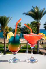 Colourful cold Rainbow Paradise and Strawberry daiquiri cocktail drinks served in glasses at pool bar overlooking blue pool, sea and palm trees, relax and holidays at sea
