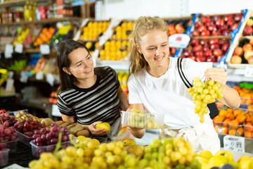 Two positive woman choosing sweet fresh grape at grocery section of supermarket