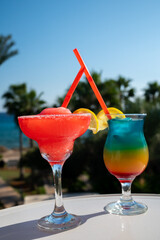 Colourful cold Strawberry daiquiri and Rainbow Paradise cocktails drinks served in glasses at outdoor cafe overlooking blue sea and palm trees, relax and holidays at sea