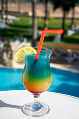 Colourful cold Rainbow Paradise cocktail drink served in glass at pool bar overlooking blue pool, sea and palm trees, relax and holidays at sea