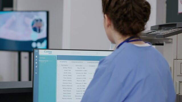 Medical nurse checking exam appointments on computer at hospital reception desk, working on checkup visits with report papers and forms. Doing healthcare insurance work in facility lobby.