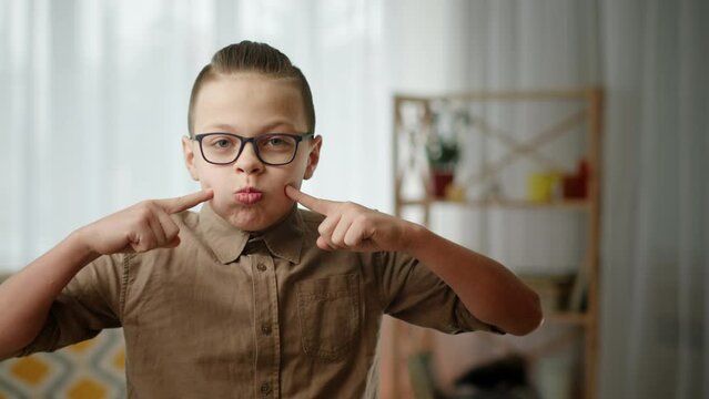 Boy in glasses poking cheeks. Funny boy in brown shirt and stylish glasses looking at camera and touching puffed cheeks while grimacing in light room at home.