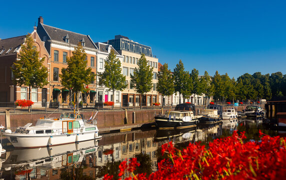 Mark River embankment in Breda, Netherlands. View of red flowers in bloom, buildings and boats along riverside.
