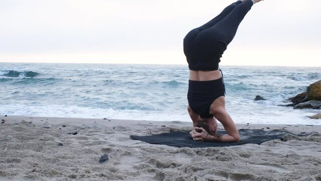 A caucasian young woman in a black sports suit doing a headstand balance asana for health on the ocean beach in a cloudy day