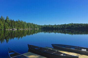 Naklejka premium Adirondack Park, New York: Canoes tied to a wooden dock on Sagamore Lake, surrounded by evergreen trees on a bright summer day.