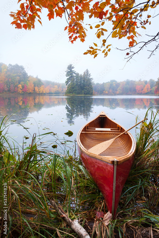 Wall mural red wooden canoe on calm lake with trees in fall color and maple branches above during autumn