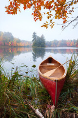 Red wooden canoe on calm lake with trees in fall color and maple branches above during autumn