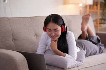 Beautiful Asian woman relaxing on sofa with laptop and wearing headphones in living room at home.